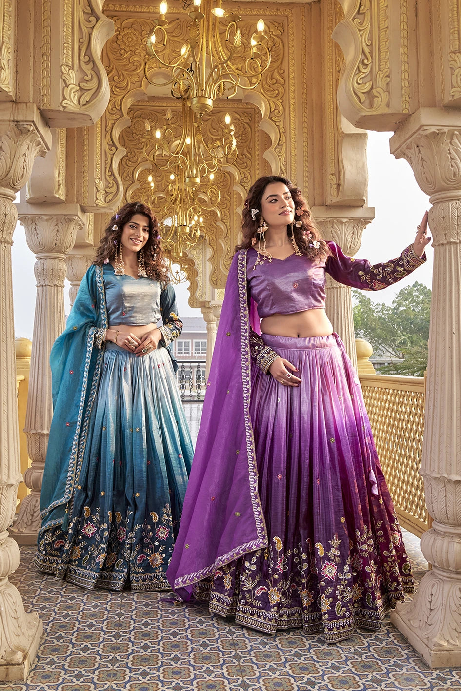 Two women in traditional blue and purple lehengas standing in an ornate architectural setting.